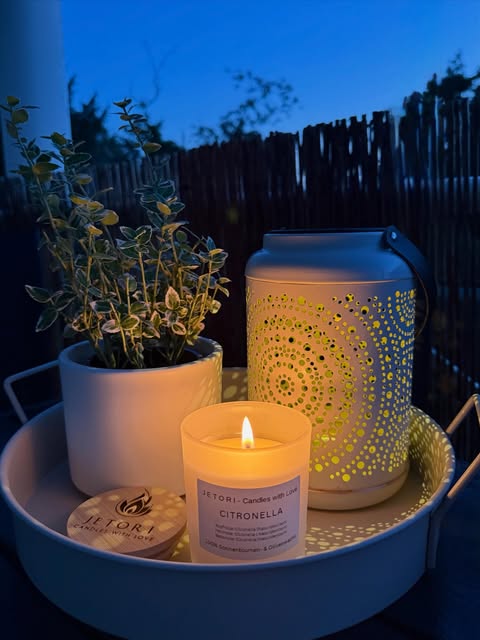 Eine leuchtende JETORI-Citronellakerze mit Holzdeckel neben einer Laterne im Abendlicht.